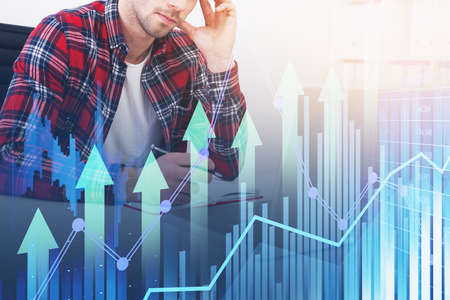 Serious young handsome businessman wearing shirt is taking notes. Workplace with desk, laptop financial graph and bar diagram in the background. Concept of work in the internet via mobile gadgetsの写真素材