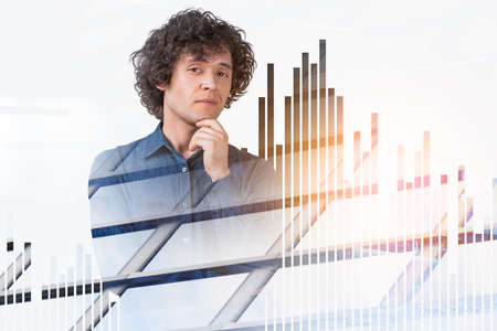 Man in denim shirt, hand to chin, pensive looking at the camera. Double exposure with office buildings and rising bar chart. Concept of business idea and investmentの写真素材