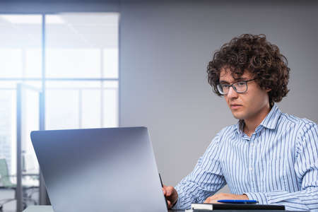 Concentrated man in shirt using laptop, notebook and office tools on desk, looking at the computer screen. Office room with windows. Concept of business secretaryの写真素材