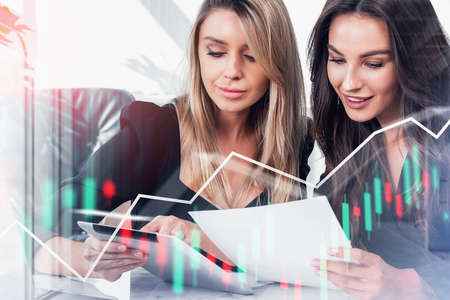 Young attractive businesswomen wearing formal suits work together talking about new profitable contract. Office workplace with forex candlesticks. Concept of teamwork, coworking and partnershipの写真素材
