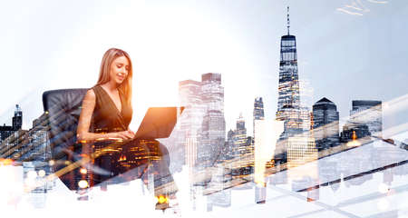 Businesswoman sitting in office armchair, working with laptop, smiling. Panoramic view of New York cityscape, toned image. Concept of finance and technologyの写真素材