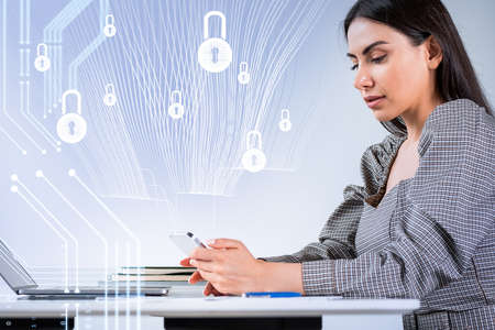 Businesswoman in formal wear works on smartphone typing message. Digital interface with icons of padlocks in the foreground. office workplace with laptop. Concept of modern technologies in businessの写真素材