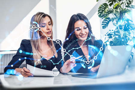 Two office women work together, laptop on desk. Team analyzing stock market changes, financial hologram. Binary blue chart with growing lines. Concept of online tradingの写真素材