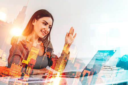 Smiling young attractive businesswoman wearing formal dress is having online video conference call via laptop. City skyscraper in the background. Concept of internet communication via gadgetsの写真素材