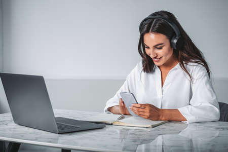 Young attractive businesswoman wearing white blouse and headphones is working on smartphone having...