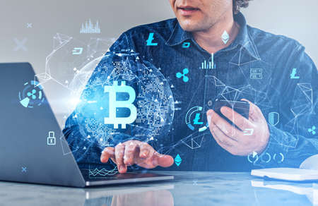Businessman wearing jeans shirt is holding smartphone and typing on laptop with digital interface with virtual globe and bitcoin cryptocurrency and diagrams. Concept of international internet tradingの写真素材