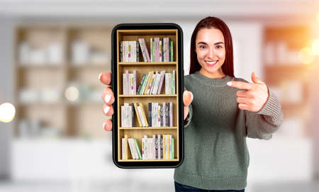 Young woman with happy look, finger point at phone with digital library in mobile app, blurred background with books on shelf. concept of online educationの写真素材