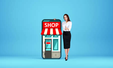Businesswoman showing thumb up, full length, standing near phone with online shop on screen, light blue background. Mockup copyspace. Concept of digital payment and buyの写真素材