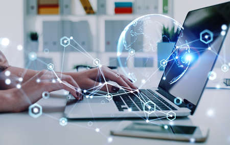 Businesswoman is typing message on laptop with hologram of virtual globe sitting at office workplace...