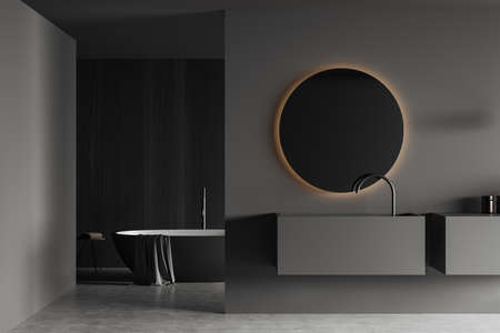 Dark bathroom interior with sink and round mirror, bathtub with towel in the corner on gray concrete...