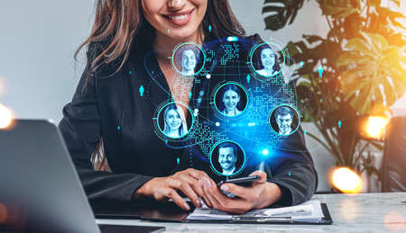 Businesswoman smiling with smartphone in hands, laptop and contract on desk. Earth globe hologram,...
