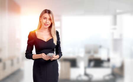Young attractive businesswoman wearing formal wear is standing holding smartphone at office...