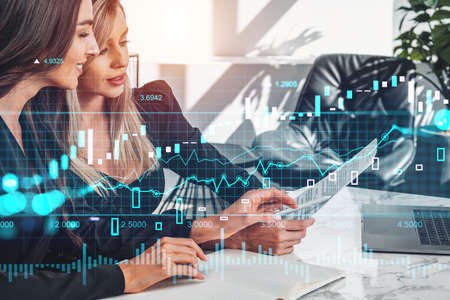Two businesswomen reading business contract, laptop on desk. Candlesticks and forex diagrams....