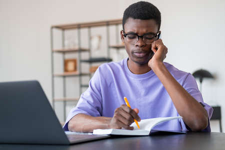 Serious black businessman call on the phone, take notes in notebook on desk with laptop. Blurred...