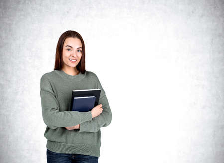 Attractive businesswoman wearing casual wear is standing holding notebook near empty white wall in...