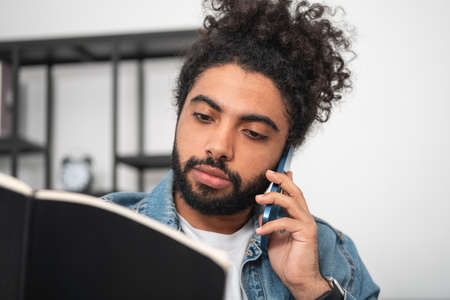 Young handsome businessman wearing casual wear is talking on smartphone and reading notes at office...