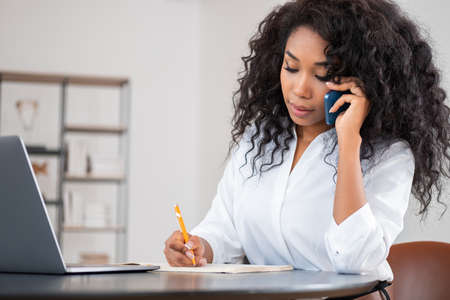 African businesswoman take note and talk on the phone, laptop and notebook on desk. Office room with shelf on blurred background. concept of communicationの写真素材