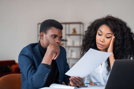 Black pensive businessman and businesswoman reading a business document together, laptop and notes...