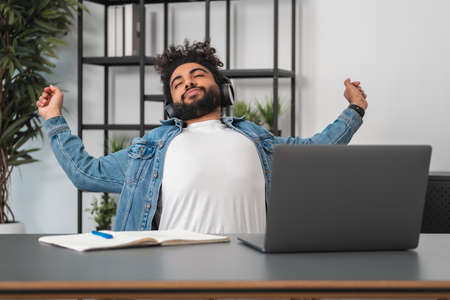 Tired handsome businessman wearing casual wear is stretching himself listening to podcast at office...