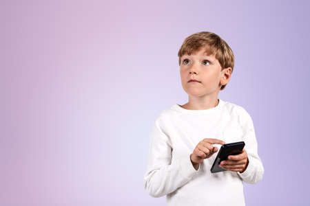 Boy child with look up finger touching smartphone, portrait on empty lilac background. Concept of social media and education. copy spaceの写真素材