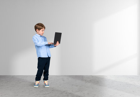 Handsome young boy wearing formal wear is standing holding laptop near empty white wall in background. Concrete floor. Concept of modern gadgets, learning, studying, progressive kidsの写真素材