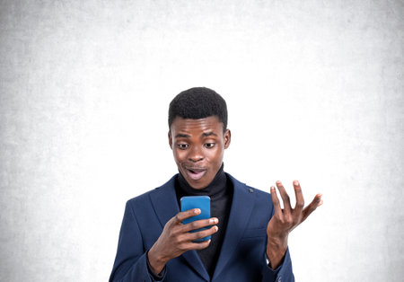 Handsome African American businessman wearing formal suit is standing watching at smartphone near empty concrete wall in background. Concept of modern gadgets, mobile communication, time managementの写真素材