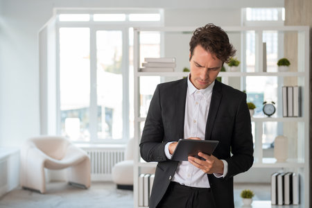 Pondering businessman wearing formal suit is standing holding tablet device and typing on gadget at office workplace. Concept of model, business person, time management, surfing net for business ideasの写真素材
