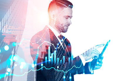 Glad handsome businessman wearing formal suit typing on tablet device with virtual hologram of forex graph, candlestick and bar diagram. city skyscrapers. Concept of successful trading on the stock marketの写真素材