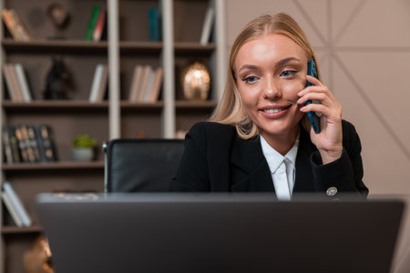 Smiling businesswoman talking on the phone, working with laptop computer. Office room with shelf and decoration on background. Concept of online network and communicationの写真素材