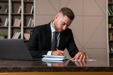 Concentrated businessman take notes sitting at the desk with laptop, shelf with decoration on...