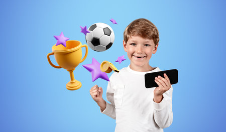 Happy child boy looking at the camera with raised fist, football and champion cup on blue...