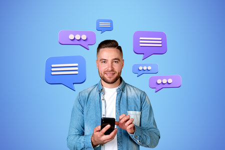 Smiling bearded young man in casual clothes using smartphone over blue background with speech...