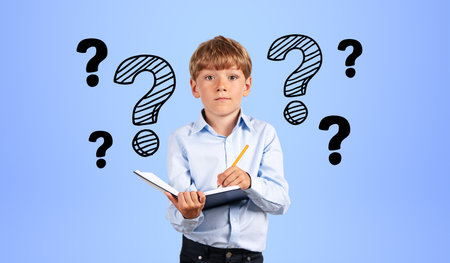 Portrait of serious little boy in blue shirt writing in notebook near blue wall with question marks drawn on it. Concept of curiosity and educationの写真素材