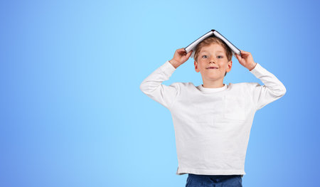 Smiling school boy with book on head, looking at the camera on copy space blue background. Concept...