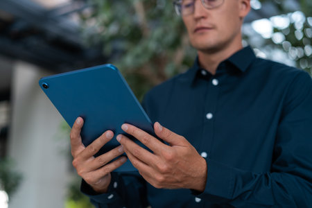 Concentrated businessman in eyeglasses looking at tablet in hands, standing low angle on blurred...