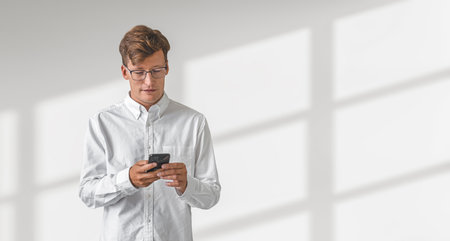 Businessman in eyeglasses typing in phone in hands, smiling portrait on shadow empty white background. Concept of social media marketing and online shoppingの写真素材