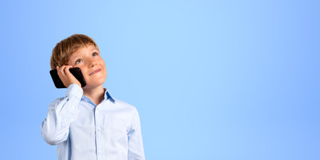 Portrait of cheerful elementary school schoolboy in formal clothes talking on smartphone and looking upwards standing over blue background. concept of communication. copy spaceの写真素材