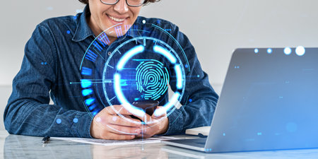 Unrecognizable smiling businessman using smartphone with HUD fingerprint interface at office table with laptop. Concept of cyber security, biometric scanning and authenticationの写真素材