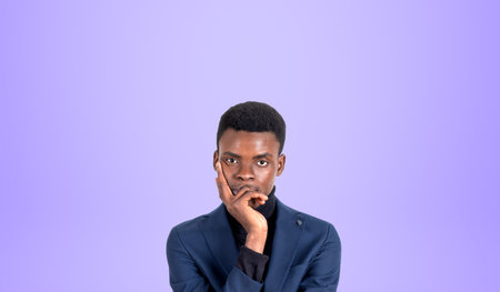 Portrait of pensive young African American businessman thinking standing with hand on chin standing over purple background. Concept of planning and contemplationの写真素材