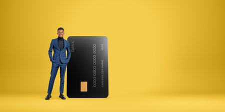 Portrait of cheerful young African American businessman standing near big credit card over orange copy space background. Concept of banking and money transactionの写真素材