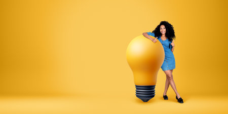 Smiling black woman showing thumb up, full length standing near large lightbulb on empty yellow...