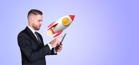 Young European businessman with smartphone standing over purple background with bitcoin rocket. Concept of cryptocurrencyの写真素材