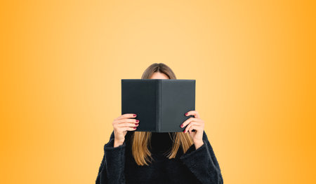 Young woman reading book hiding face, student holding textbook or carefully studying on empty orange...