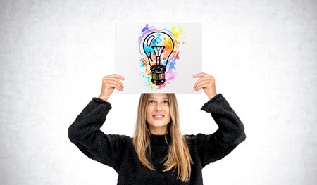 Smiling young woman showing poster with colorful lightbulb above head, gray concrete wall background. Concept of inspiration, start up, creativity, knowledge and ideaの写真素材