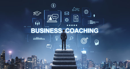 A businessman stands on stairs, gazing at hologram icons and the text Business Coaching. The background is a cityscape at night, conveying the concept of business coaching and future successの写真素材