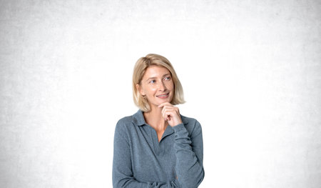 Smiling and thoughtful blonde woman with hand on chin, portrait looking aside on copy space gray concrete wall background. Making up plan and search for answers. Concept of idea or strategyの写真素材