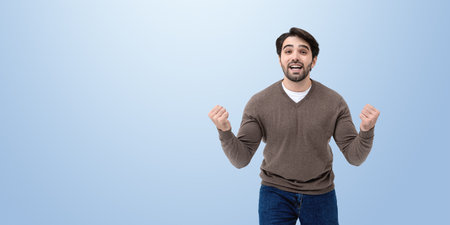 Excited young man with beard and fists up, happy winner on empty copy space blue background. Concept of celebration of success, profit, victory and luckの写真素材