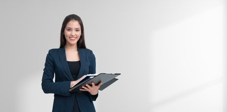 A smiling businesswoman in formal attire holds a clipboard. She stands confidently against a light background, symbolizing professionalism and successの写真素材