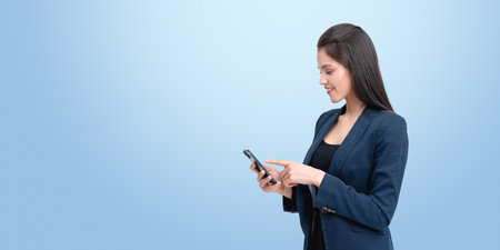 Smiling woman in business attire using a smartphone, standing against a light blue background, representing technology and communicationの写真素材