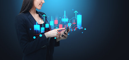 Businesswoman in dark suit using smartphone, analyzing stock data on digital screen. Dark blue background, concept of finance and technologyの写真素材
